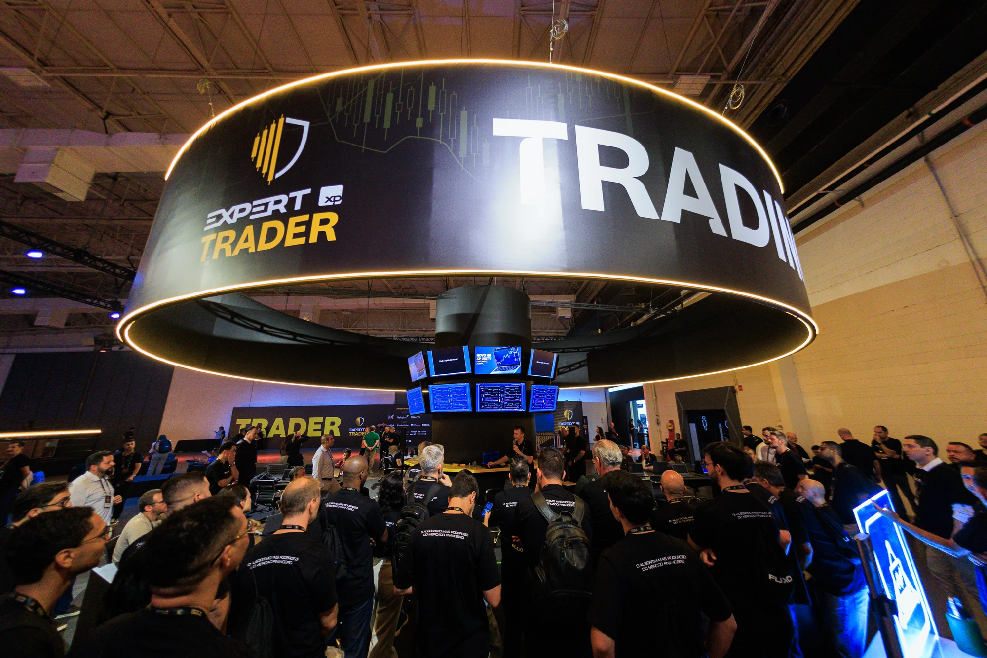 Trading Lab visto por inteiro, com anel luminoso suspenso e mesas ocupadas pelo público