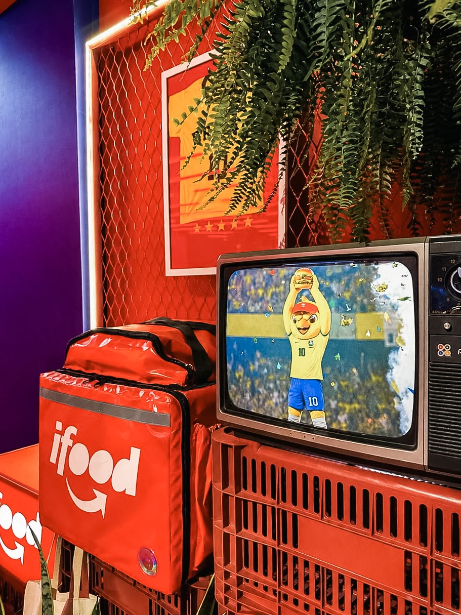 Detalhe da bag do iFood ao lado de uma televisão com o Canarinho